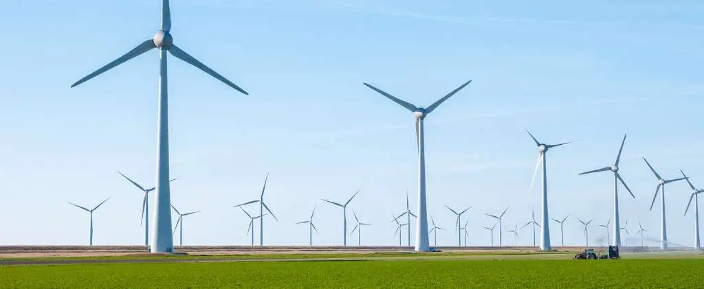 Un champ avec de nombreuses éoliennes produisant de l'électricité sous un ciel bleu clair, illustrant les solutions énergétiques de premier ordre. Un tracteur est visible au premier plan sur l'herbe verte.