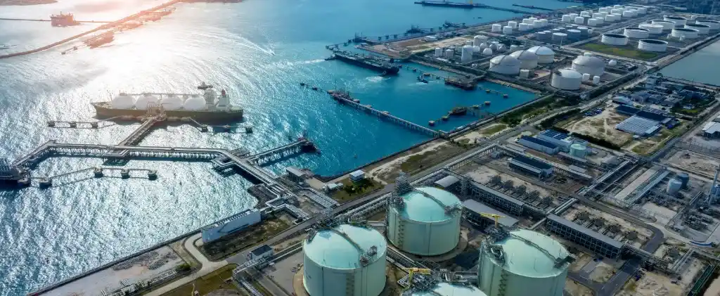 Vue aérienne d'une installation industrielle côtière, animée par des activités d'importation et comportant des réservoirs de stockage, des pipelines et des quais par une journée ensoleillée. Les navires liés au commerce du gaz sont amarrés aux quais, tandis que de nombreux grands réservoirs blancs dominent le site.