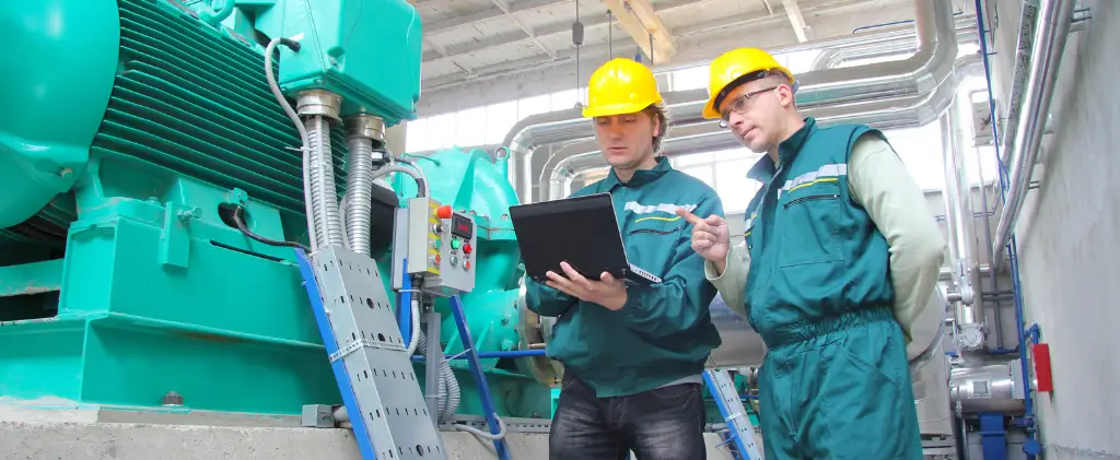 Deux ouvriers en uniformes verts et casques jaunes regardent un ordinateur portable dans un environnement industriel, inspectant l'équipement avec une attention méticuleuse à la capacité d'effacement de la machine.