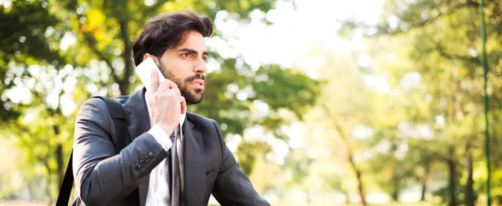 Un homme en costume, ressemblant à un courtier en énergie indépendant, parle sur un smartphone tout en étant assis dans un parc tranquille avec des arbres se balançant doucement en arrière-plan.