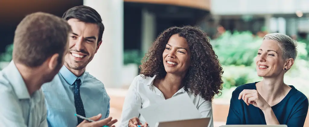 Quatre personnes sont assises ensemble à une table dans un cadre intérieur lumineux, engagées dans une conversation animée sur les moyens d'économiser l'énergie en entreprise et souriant tout en partageant des idées.