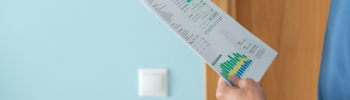 Personne tenant des documents imprimés contenant des graphiques et des données, debout devant une porte en bois et un mur bleu clair avec un interrupteur, examinant éventuellement des statistiques liées aux réacteurs nucléaires.