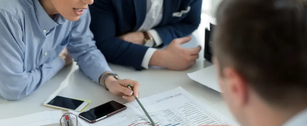 Trois personnes sont réunies autour d'une table, discutant d'un document avec des plans architecturaux. Une personne montre les plans avec un stylo, soulignant éventuellement des aspects liés à son contrat énergie pro. Divers appareils et papiers sont également sur la table.