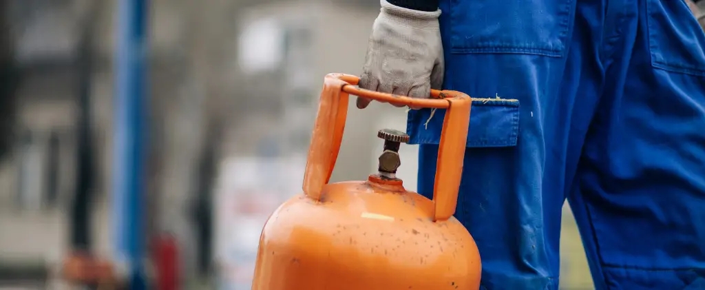 Travailleur en combinaison bleue tenant une bouteille de gaz orange par sa poignée avec une main gantée, assurant la sécurité de son contrat énergie pro.
