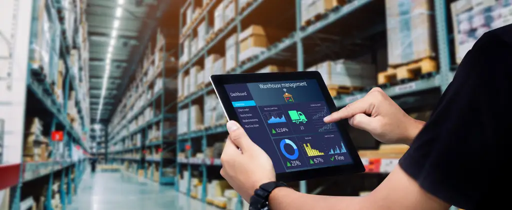Personne utilisant une tablette pour gérer les stocks dans un grand entrepôt avec des étagères remplies de cartons. L'écran de la tablette affiche des analyses liées à la gestion de l'entrepôt et de l'énergie.