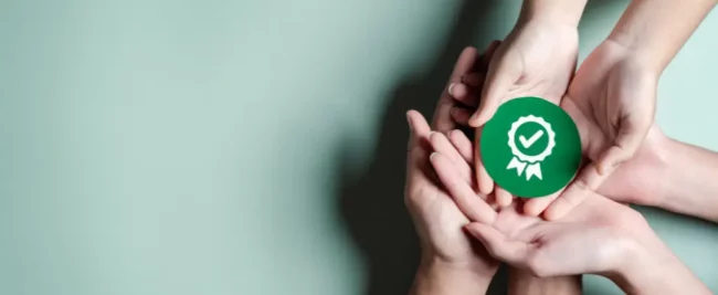 Plusieurs mains tiennent un badge circulaire vert avec une coche blanche et un symbole en ruban, signifiant l'approbation ou la certification selon la norme ISO 50001, sur fond vert clair.