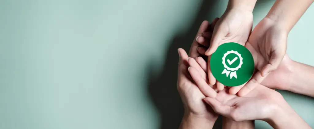 Plusieurs mains tiennent un badge circulaire vert avec une coche blanche et un symbole en ruban, signifiant l'approbation ou la certification selon la norme ISO 50001, sur fond vert clair.