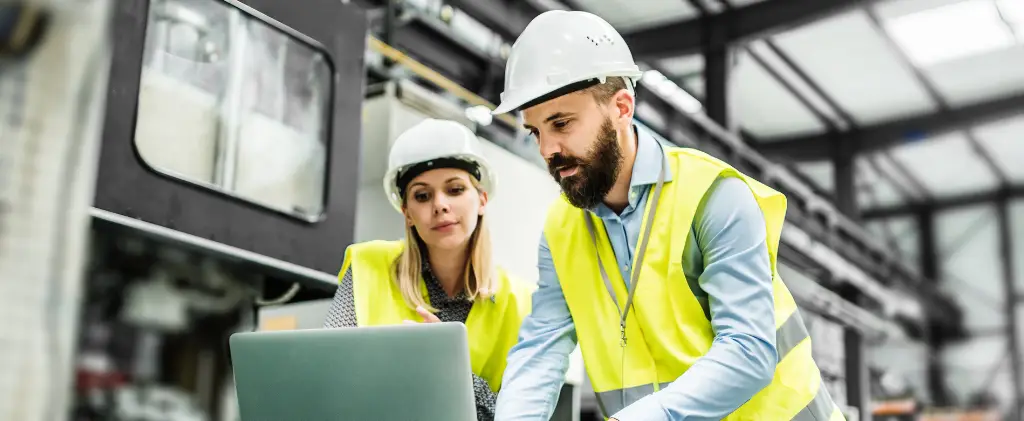 Deux travailleurs portant des casques de sécurité et des gilets de haute visibilité examinent les informations sur un ordinateur portable dans un environnement industriel, garantissant ainsi la plus grande efficacité énergétique industrielle.