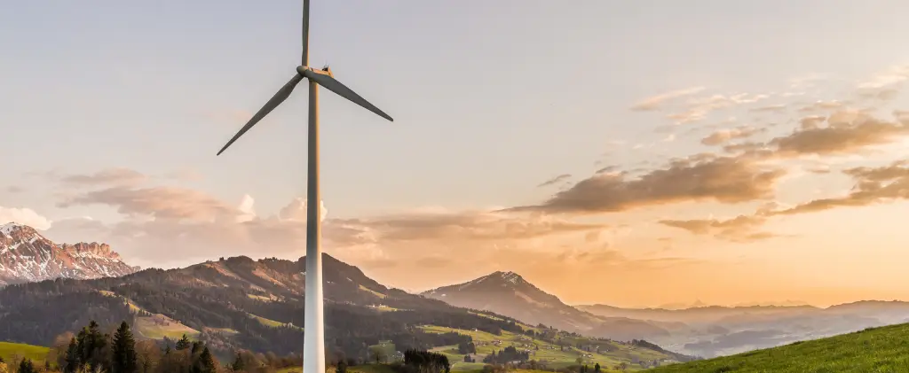 Une seule éolienne se dresse sur une colline herbeuse avec un paysage montagneux en arrière-plan au coucher du soleil, symbolisant la transition écologique et énergétique.
