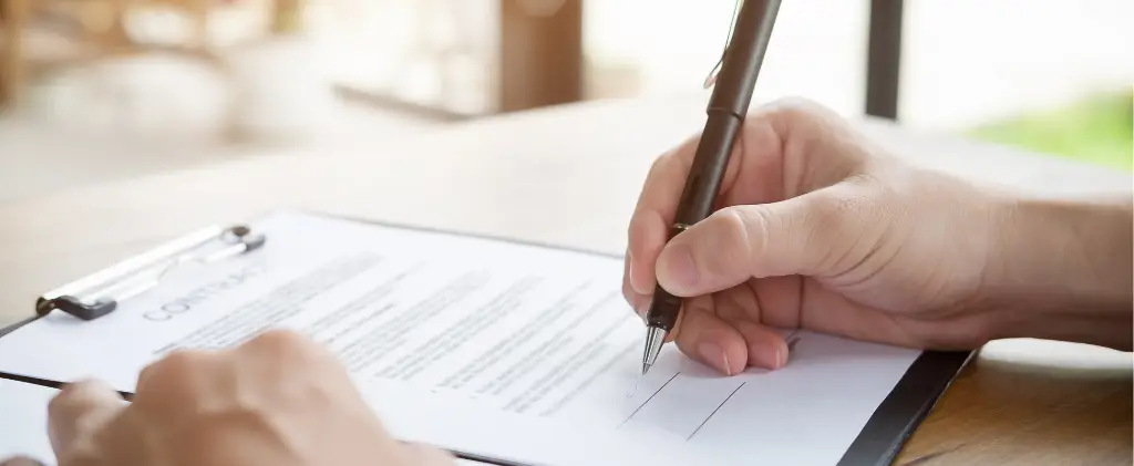 Une personne signe un document sur un presse-papiers avec un stylo sur une table en bois.
