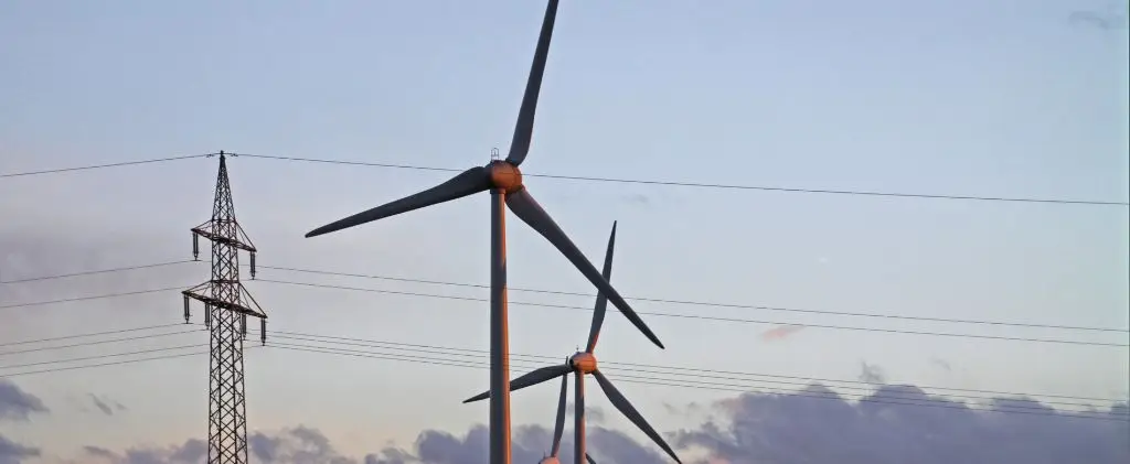 Deux éoliennes et un pylône de ligne électrique se détachent sous un ciel nuageux au crépuscule, représentant la vision avant-gardiste d'un fournisseur d'électricité professionnel en 2025.