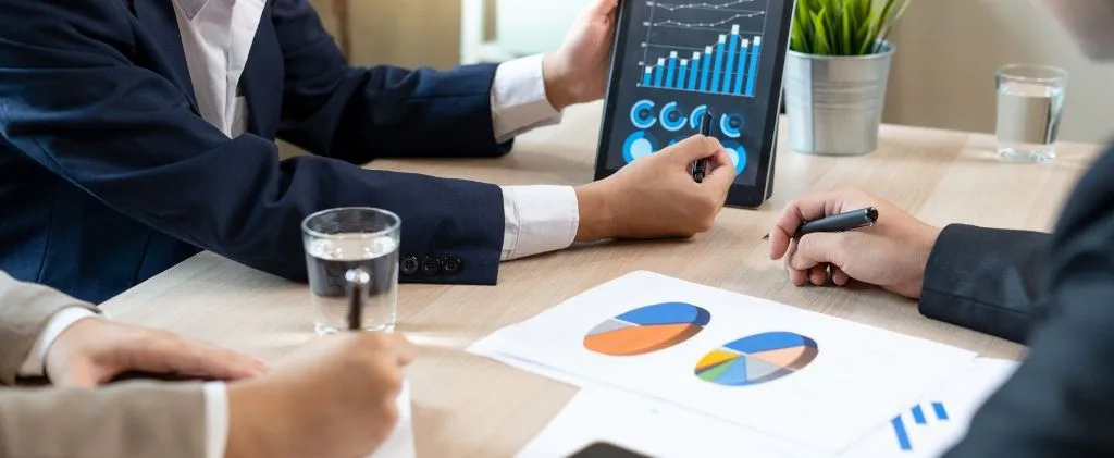 Autour d'une table de réunion, des professionnels de CEE entreprises en tenue professionnelle discutent de tableaux et de graphiques sur une tablette et des documents.