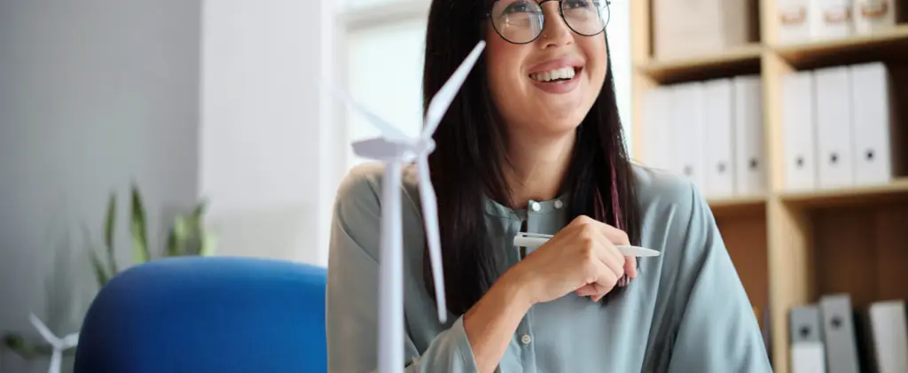 Une femme à son bureau sourit tout en tenant un stylo, représentant son expertise en conseil en énergie. Devant elle se trouve une petite maquette d'éolienne, symbole d'innovation et de durabilité.