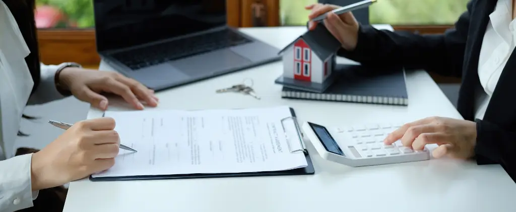Deux personnes assises à un bureau avec un contrat, un ordinateur portable, une maquette de maison et une calculatrice discutent de questions immobilières ou financières. Des clés et un contrat énergie pro reposent sur la table, laissant entrevoir des accords énergétiques entremêlés à leurs négociations.