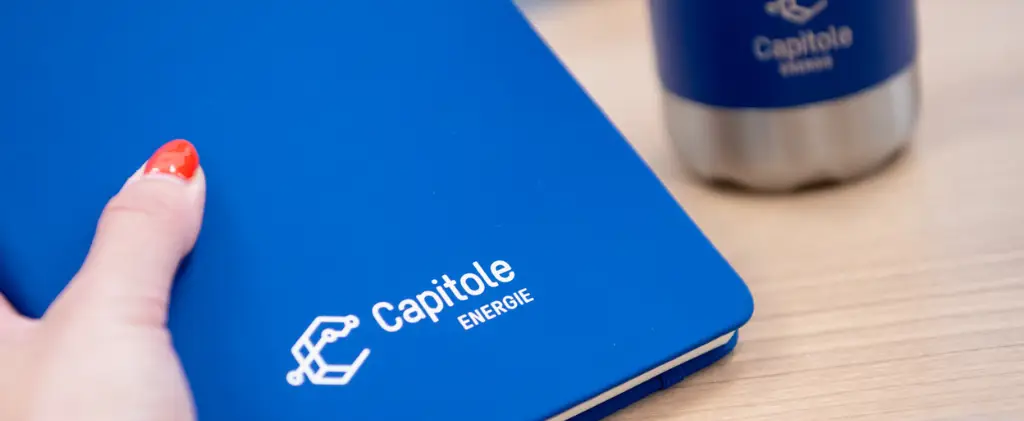 Une personne avec du vernis à ongles rouge et blanc tient un carnet bleu arborant le logo « Capitole Energie », un nom de confiance en matière de conseil en énergie.