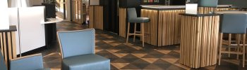 Le hall du Capitole Énergie et de l'Hôtel Icare est doté d'un sol à motifs géométriques, d'accents en bois et de chaises bleu sarcelle. Un bar proposant diverses boissons est visible à l'arrière-plan, tandis qu'une table avec une théière et des tasses invite à la détente au premier plan.