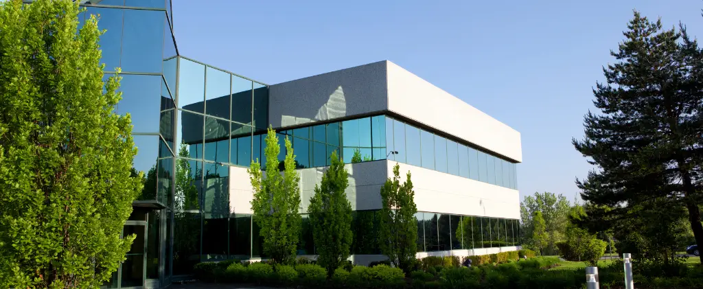 L'immeuble de bureaux moderne en verre, réputé pour son efficacité énergétique, est niché au milieu d'arbres et d'une verdure luxuriante sous un ciel bleu clair.