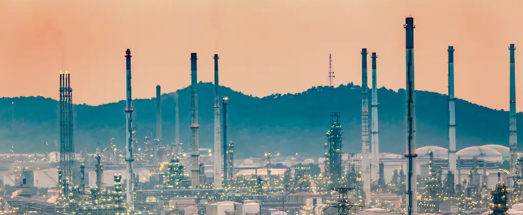 Un paysage industriel avec de nombreuses cheminées et cheminées se détachant sur un ciel brumeux au coucher du soleil, où les opérations d'un fournisseur de gaz industriel bourdonnent sans interruption. En arrière-plan, des montagnes majestueuses encadrent cette scène puissante.