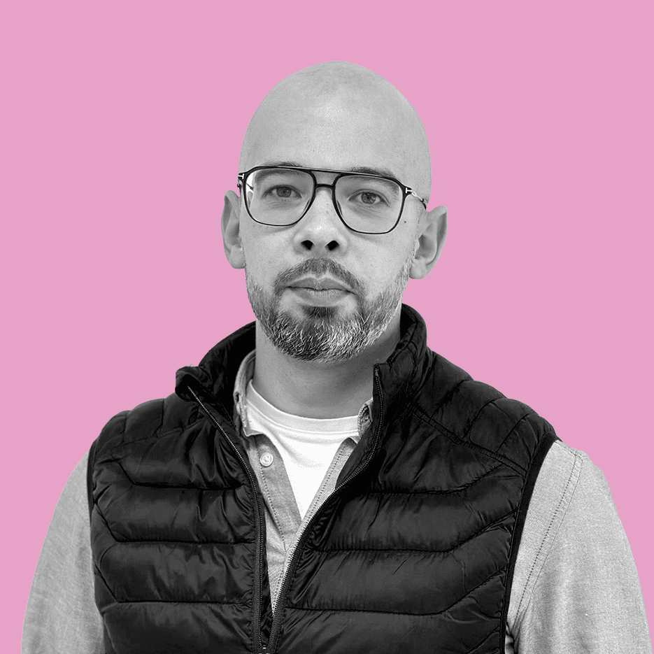 Un homme chauve avec des lunettes, représentant les solutions d’excellence, vêtu d’un gilet noir et d’une chemise grise, se tient debout sur un fond rose vif.