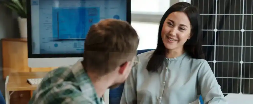 Deux personnes discutent dans un bureau, où un écran d'ordinateur affiche des dessins techniques à côté d'un panneau solaire, soulignant leur volonté de réduire la consommation d'énergie des bureaux.