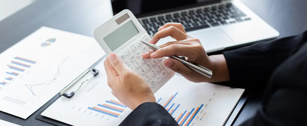 Une personne utilisant une calculatrice et tenant un stylo survole des documents financiers avec des graphiques, suivant l'évolution des accises 2025, près d'un ordinateur portable ouvert sur un bureau.