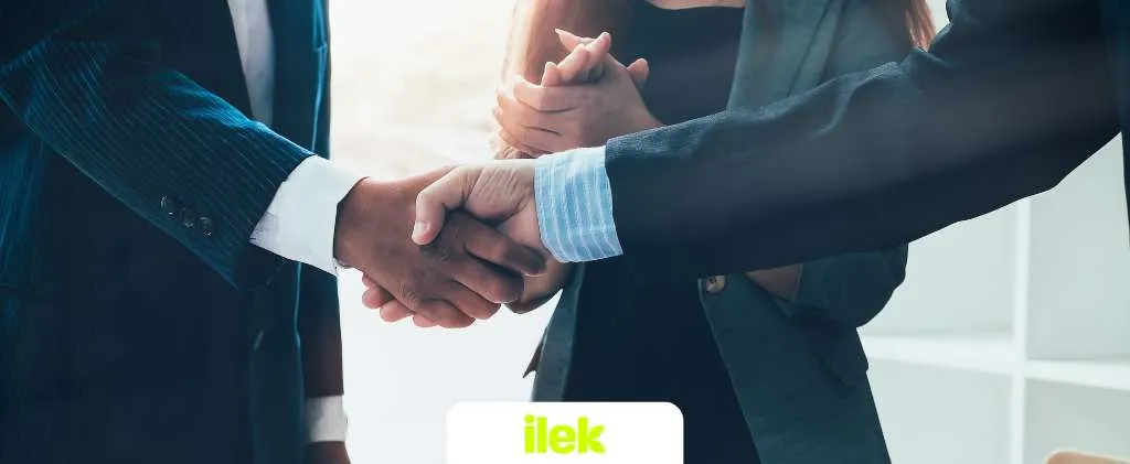 Lors d'une réunion professionnelle, trois personnes en tenue professionnelle concluent un accord par de chaleureuses poignées de main. Au premier plan, deux mains sont fermement jointes tandis qu'une femme, représentant la direction d'Ilek Pro, se tient debout, les mains soigneusement jointes.