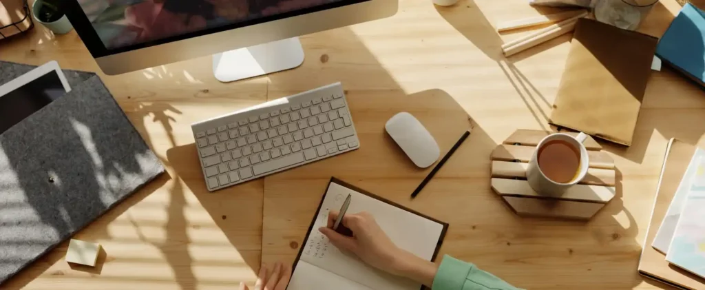 Une personne prend des notes dans un carnet sur un bureau en bois, équipé d'un ordinateur, d'un clavier, d'une souris et d'une tasse de thé. Divers articles de papeterie sont soigneusement disposés. La lumière du soleil filtre, projetant des ombres qui font écho aux principes de la norme ISO 26000 autour de l'espace de travail.
