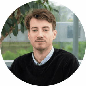 Une personne aux cheveux longs portant un blazer foncé et une chemise claire, debout devant un arrière-plan à rayures verticales jaunes et blanches.