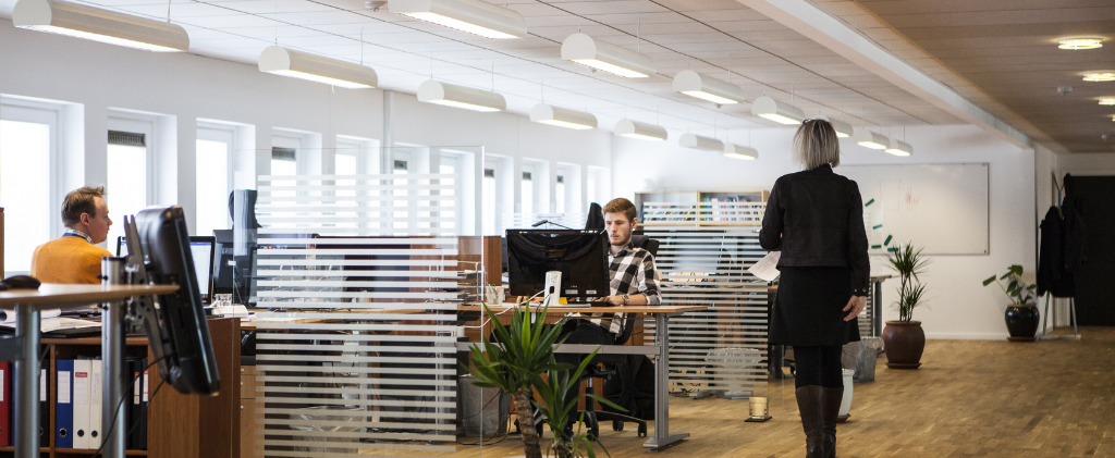 Bureau ouvert avec plusieurs personnes travaillant à des bureaux, des cloisons en verre, des plantes d'intérieur et une personne marchant le long de l'allée - un espace idéal pour les équipes qui gèrent efficacement un contrat multisite énergie.
