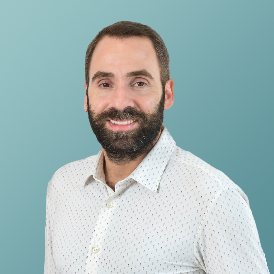 Un homme barbu aux cheveux courts sourit à la caméra, vêtu d'une chemise à motifs de couleur claire. Le fond bleu canard reflète l'ambiance sereine d'une étude bilan carbone entreprise, reflétant son engagement envers des pratiques commerciales durables.