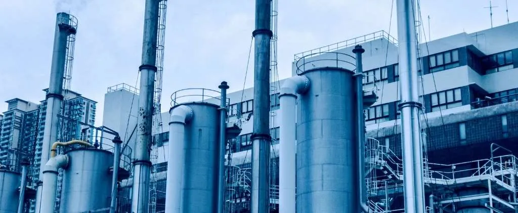 Installation industrielle avec plusieurs hautes cheminées et tuyaux métalliques cylindriques sur fond de paysage urbain.