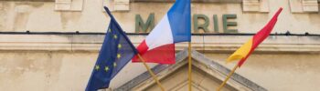 Les drapeaux de l'Union européenne, de la France et de l'Occitanie flottent fièrement devant la mairie. À proximité, un véhicule de la collectivité Enedis ronronne doucement, soulignant l'harmonie entre tradition et modernité des infrastructures.