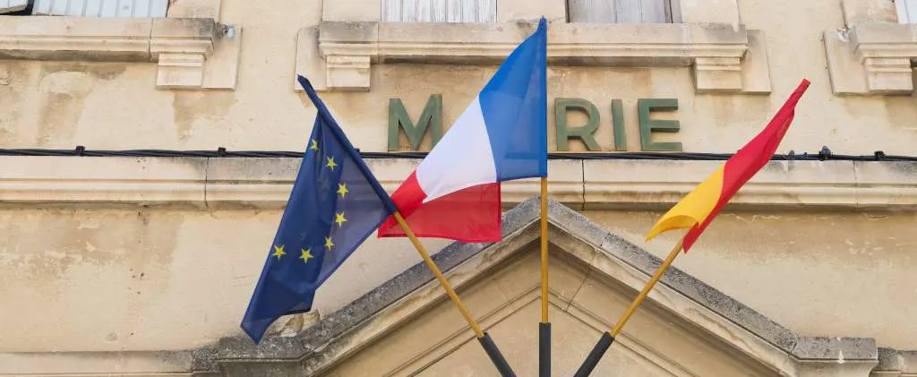 Les drapeaux de l'Union européenne, de la France et de l'Occitanie flottent fièrement devant la mairie. À proximité, un véhicule de la collectivité Enedis ronronne doucement, soulignant l'harmonie entre tradition et modernité des infrastructures.