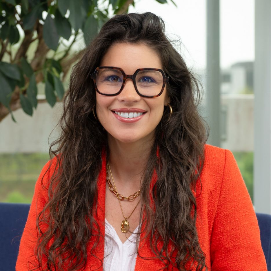 Une femme aux longs cheveux bruns ondulés, portant de grandes lunettes, un blazer rouge et des bijoux en or, est assise à l'intérieur près d'une plante et d'une grande fenêtre dans ce qui semble être une bibliothèque confortable, souriant à la caméra.
