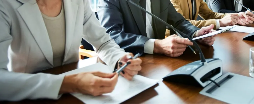 Plusieurs personnes sont assises à une table, munies de microphones, lors d'une réunion, et discutent des implications de leurs projets récents. Une personne prend des notes sur un bloc-notes.