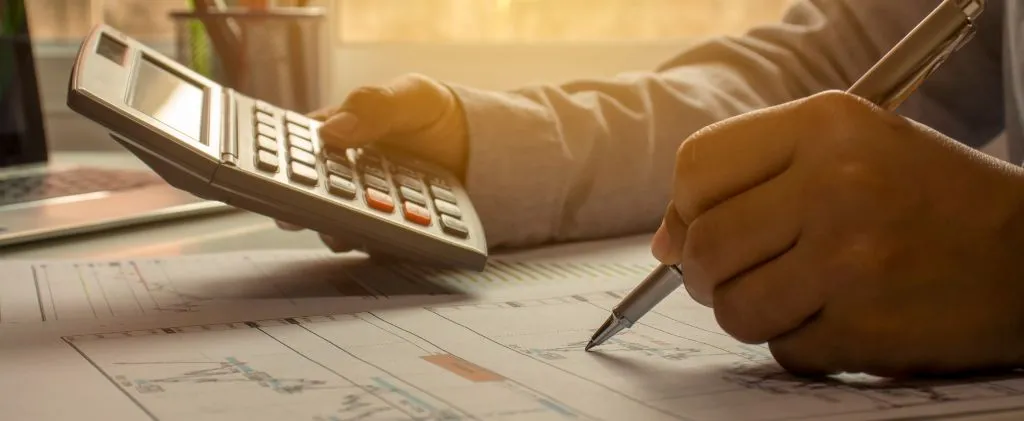 Une personne se concentre sur le calcul de chiffres sur un plan avec un stylo et une calculatrice, entourée d'un ordinateur portable et de papeterie.