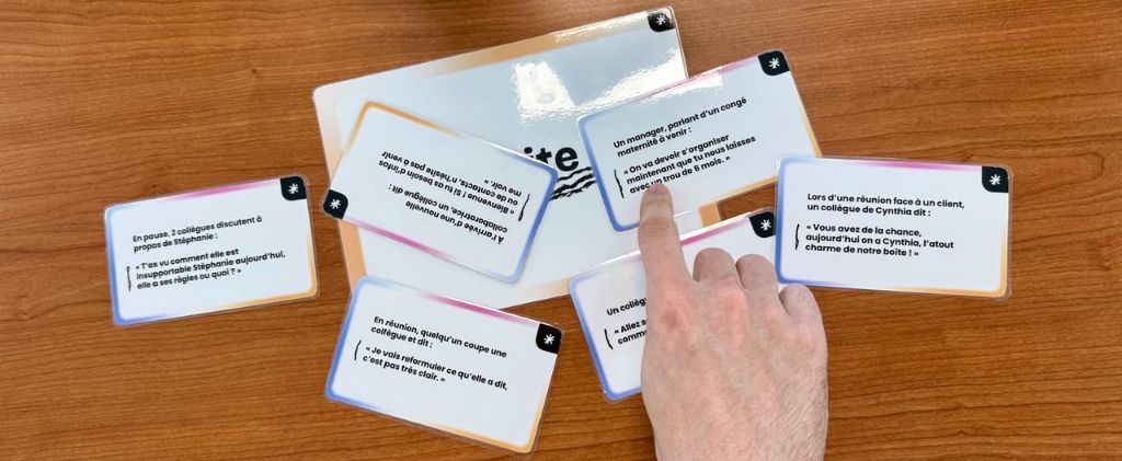 Une main désigne l'une des nombreuses cartes de sensibilisation plastifiées, avec texte en français, étalées sur une table en bois ; il s'agit probablement d'outils de sensibilisation au harcèlement. Un panneau partiellement visible se trouve sous les cartes.