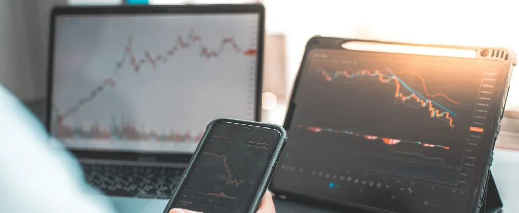 Une personne tient un smartphone affichant un graphique boursier sur Capn, avec un ordinateur portable et une tablette en arrière-plan affichant des graphiques financiers et des données de marché.