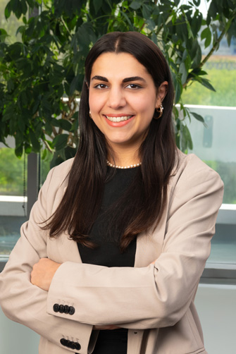 Une femme aux longs cheveux noirs, vêtue d'un blazer beige et d'un haut noir, se tient à l'intérieur, les bras croisés, souriant devant une plante feuillue et une fenêtre, rayonnant d'énergie et de confiance comme si elle était prête à écrire sa propre histoire.