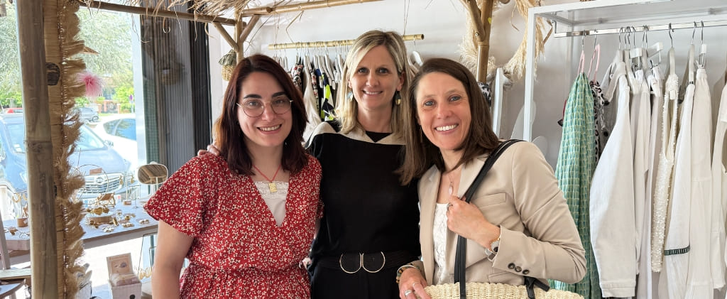 Trois femmes se tiennent ensemble et sourient dans une boutique, avec des vêtements et des accessoires visibles en arrière-plan, après avoir visité Capitole Énergie et La Fromagerie des Cabasses.