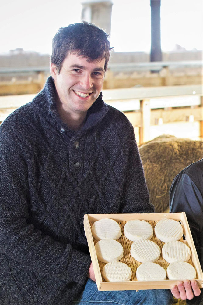 Une personne portant un pull foncé tient un plateau en bois avec neuf morceaux ronds de fromage blanc dans un décor intérieur.