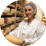 Une personne portant une blouse blanche et un filet à cheveux tient une meule de fromage dans une salle de stockage remplie d’étagères de meules de fromage en cours de vieillissement — une image qui rappelle l’Étude de cas : Fromagerie de Cabasses et Capitole Énergie.