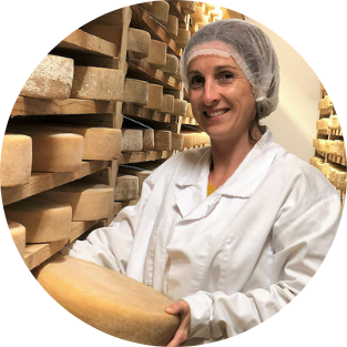 Une personne portant une blouse blanche et un filet à cheveux tient une meule de fromage dans une salle de stockage remplie d’étagères de meules de fromage en cours de vieillissement — une image qui rappelle l’Étude de cas : Fromagerie de Cabasses et Capitole Énergie.