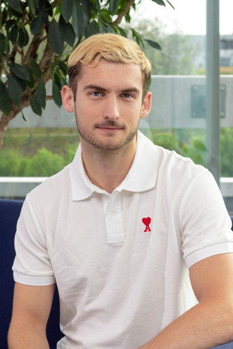 Un jeune homme aux cheveux clairs et à la barbe courte, portant un polo blanc avec un cœur rouge et un logo en ruban, est assis à l'intérieur près d'une fenêtre et d'une plante feuillue, reflétant l'énergie capitole de son histoire inspirante.