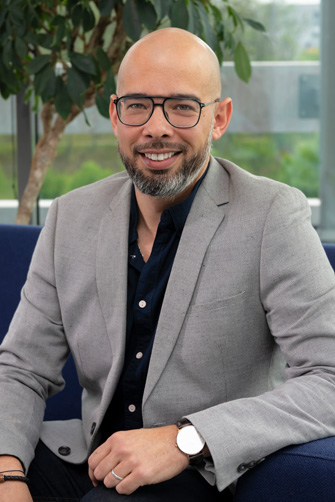 Un homme avec des lunettes et une barbe, vêtu d'un blazer gris et d'une chemise sombre, est assis sur un canapé bleu devant une fenêtre avec de la verdure en arrière-plan, réfléchissant à l'histoire du Capitole et à son énergie.