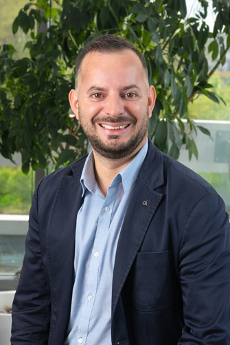 Un homme aux cheveux courts et à la barbe, vêtu d'une chemise bleue et d'un blazer foncé, sourit assis à l'intérieur devant une grande plante verte, l'énergie capitole de sa présence ajoutant à l'atmosphère vibrante et laissant entrevoir une histoire unique.