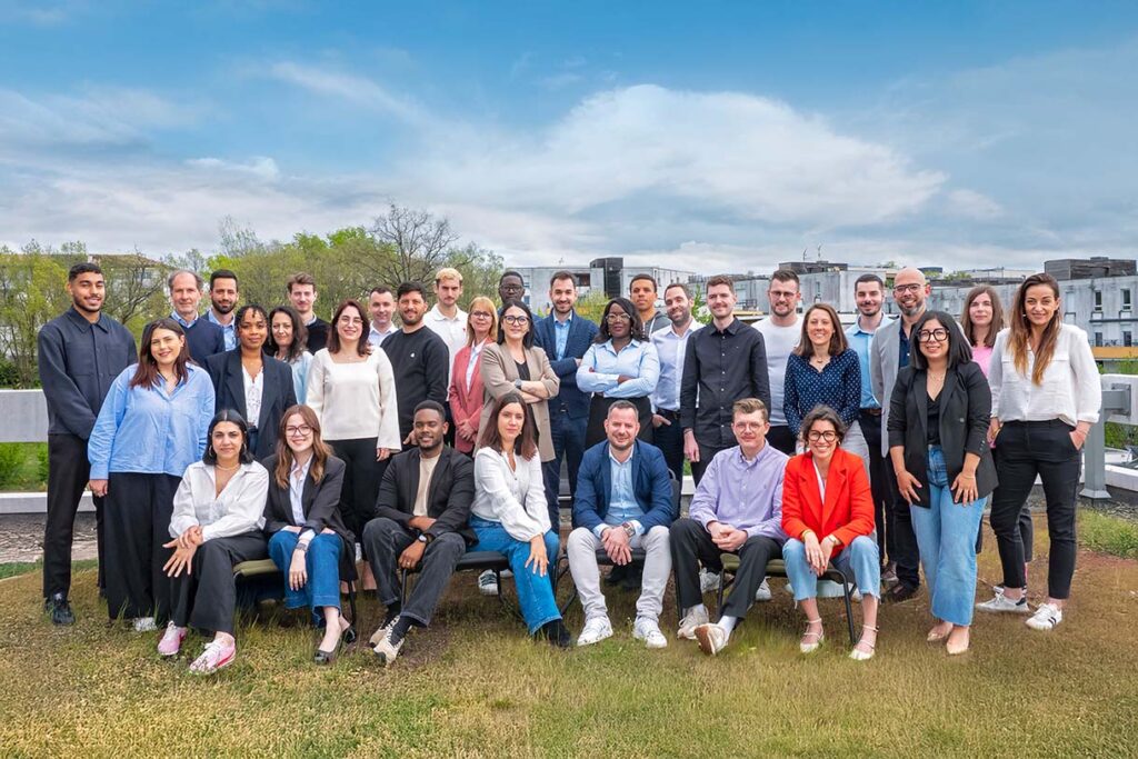Un groupe d'environ 35 personnes, dont des professionnels de Courtier en énergie entreprises, posent ensemble à l'extérieur sur l'herbe avec des bâtiments et des arbres en arrière-plan sous un ciel partiellement nuageux.