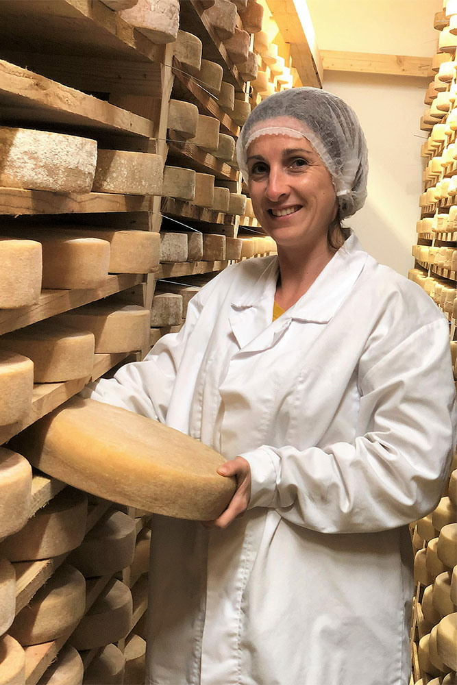 Une personne vêtue d'une blouse blanche et d'un filet à cheveux tient une grande meule de fromage dans une salle de stockage remplie d'étagères de meules de fromage vieillissantes.