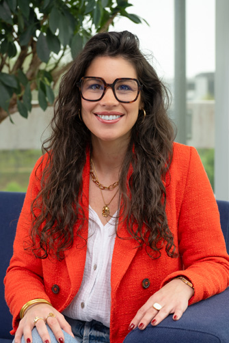 Une femme aux cheveux longs et ondulés et portant de grandes lunettes est assise sur une chaise bleue, portant un blazer orange vif qui rayonne d'énergie, un haut blanc et des bijoux en or, souriant à la caméra.