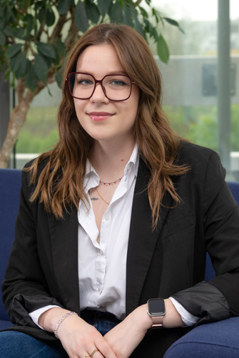 Une jeune femme aux longs cheveux bruns, portant des lunettes, un blazer noir, une chemise blanche et une montre connectée, est assise sur une chaise bleue à l'intérieur avec de la verdure en arrière-plan, dégageant l'énergie et la confiance du Capitole.